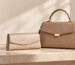 Taupe top-handle handbag beside a matching evening purse on a neutral stone surface